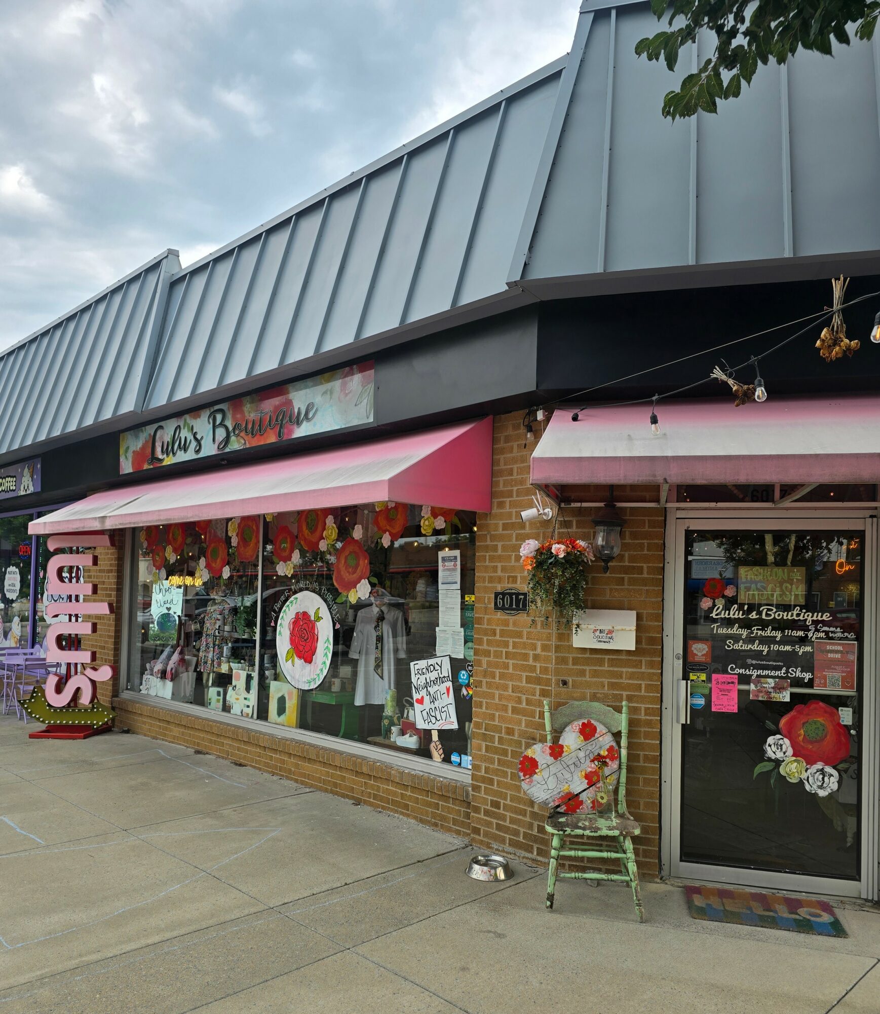 Storefront with pink awning, decorated window display, and a chair with a floral cushion outside, under a cloudy sky.