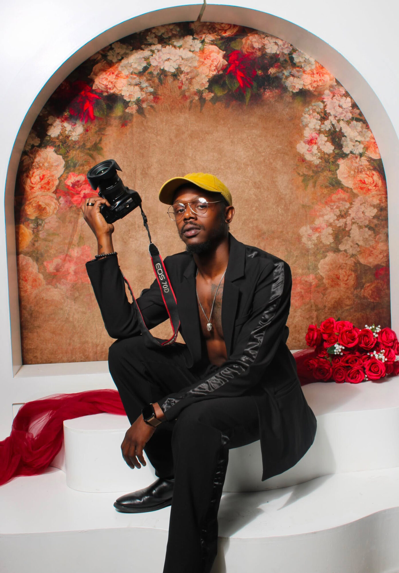 Person sitting on a white bench holding a camera, wearing a black suit and yellow cap, with floral background.