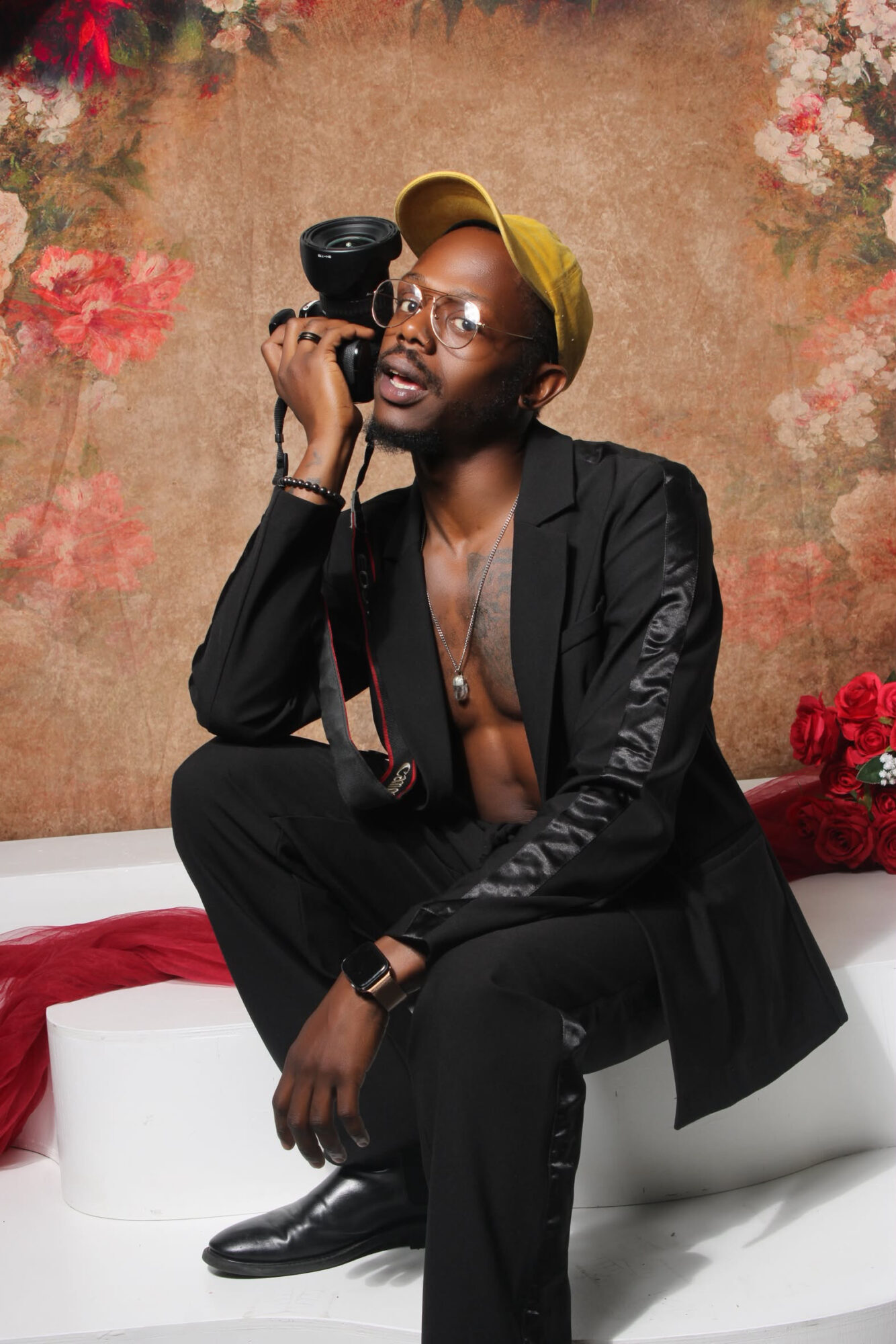 Person sitting on a white surface, holding a camera to their face, wearing glasses, a yellow headband, and a black blazer, with a floral background.