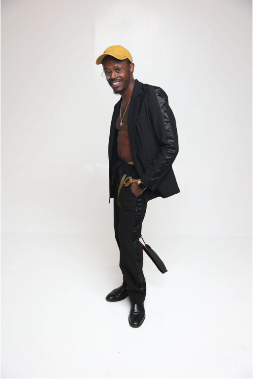 Person standing, smiling, wearing a yellow cap, black jacket, black pants, and black shoes, against a plain white background.