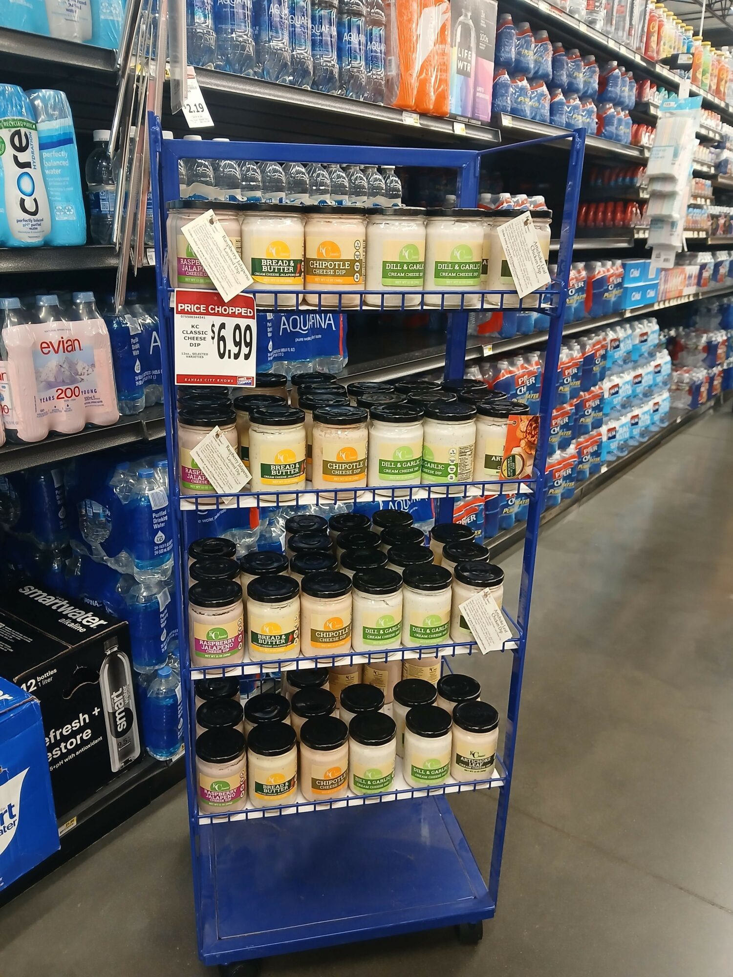 Blue display rack with various bottles and containers, store shelves in background, shopping cart handle visible, store interior.