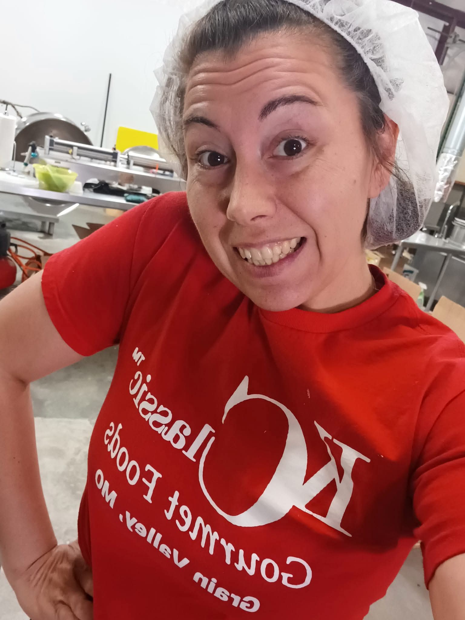 Woman in red shirt smiling in a kitchen or food preparation area, wearing a hairnet.