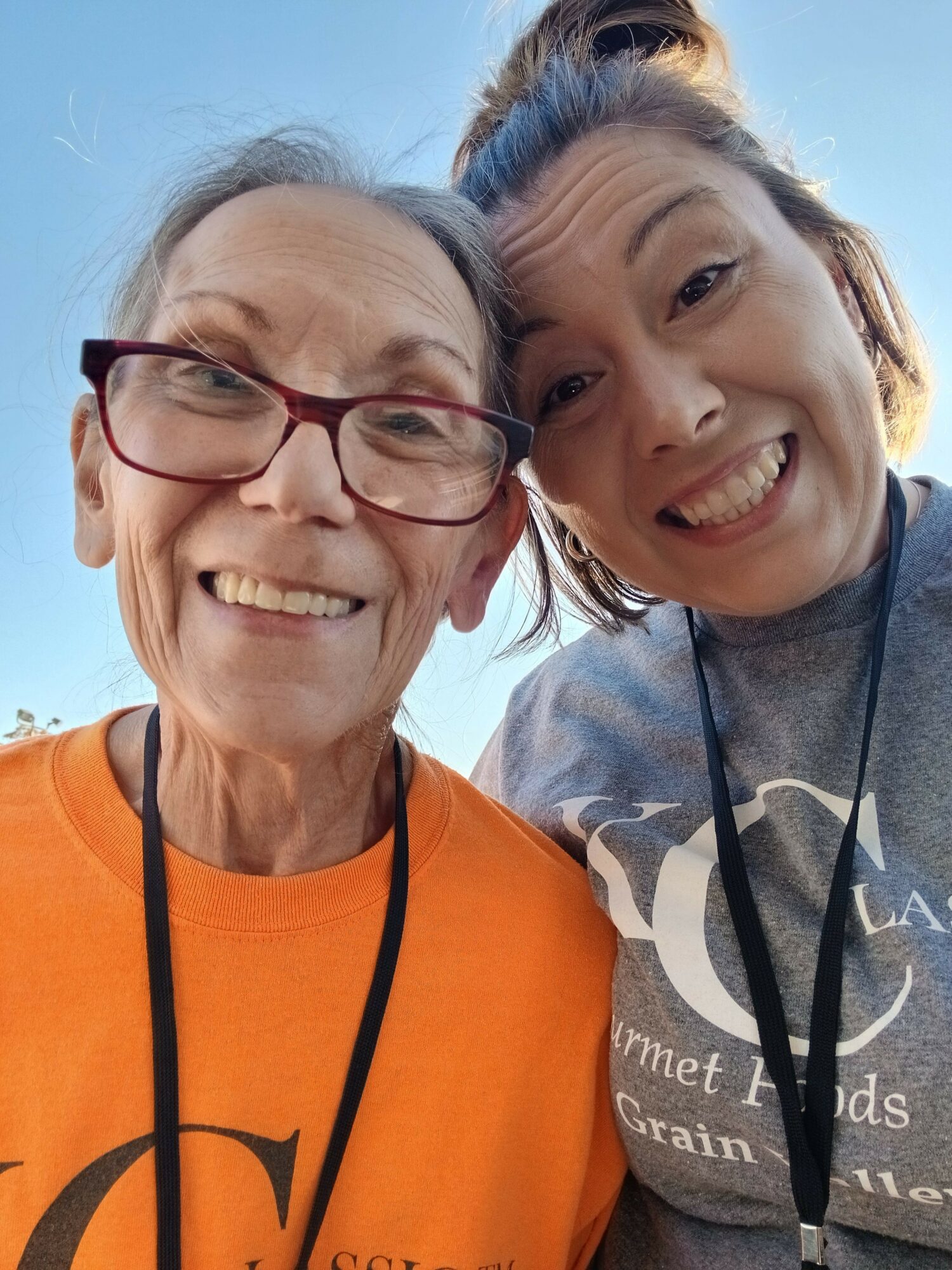 Two smiling women taking a selfie outdoors, one wearing glasses and an orange shirt, the other with a gray shirt, both smiling.