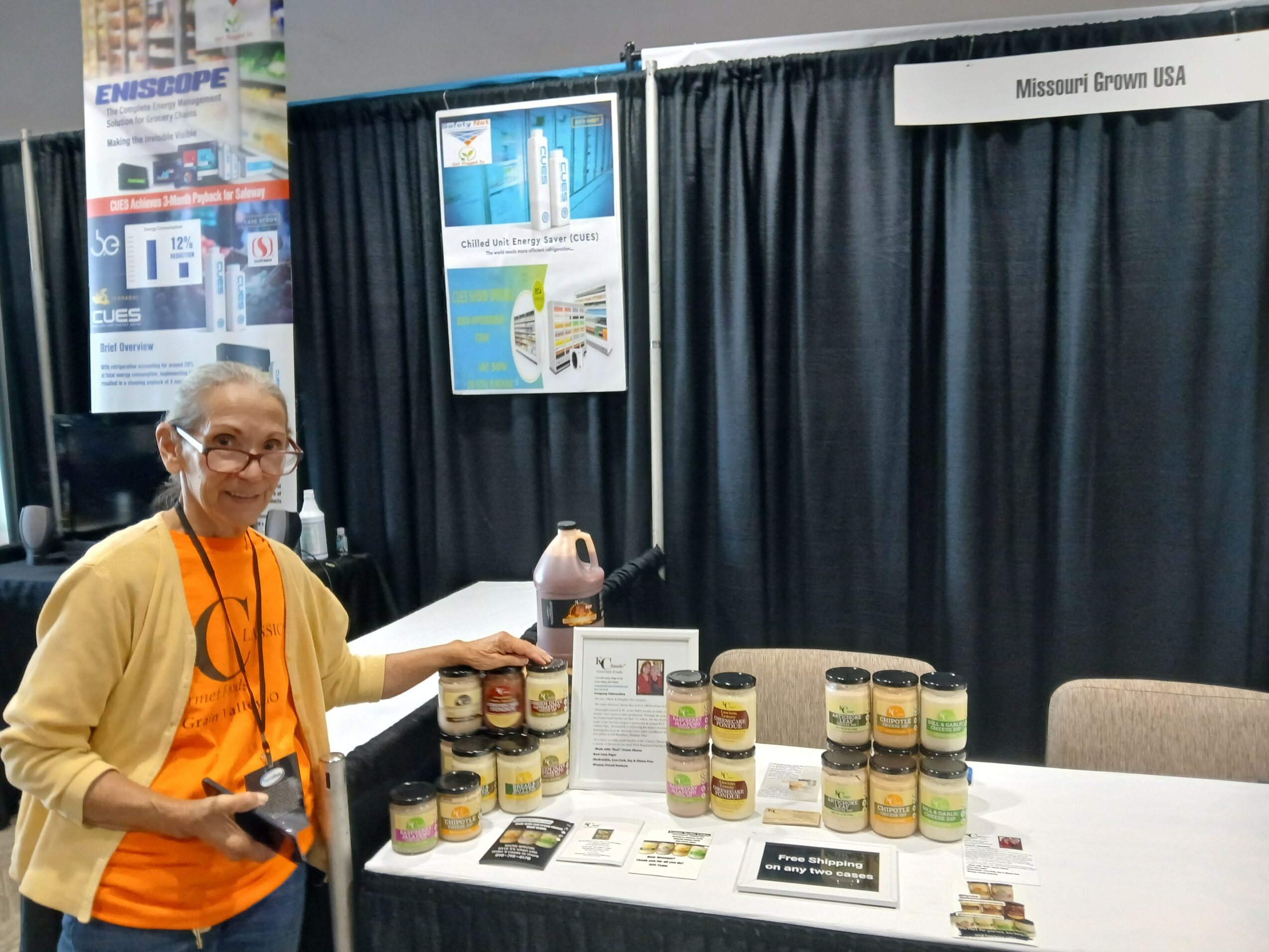 Woman in orange shirt and yellow cardigan standing next to a display of jars at an exhibition booth, black curtains behind.