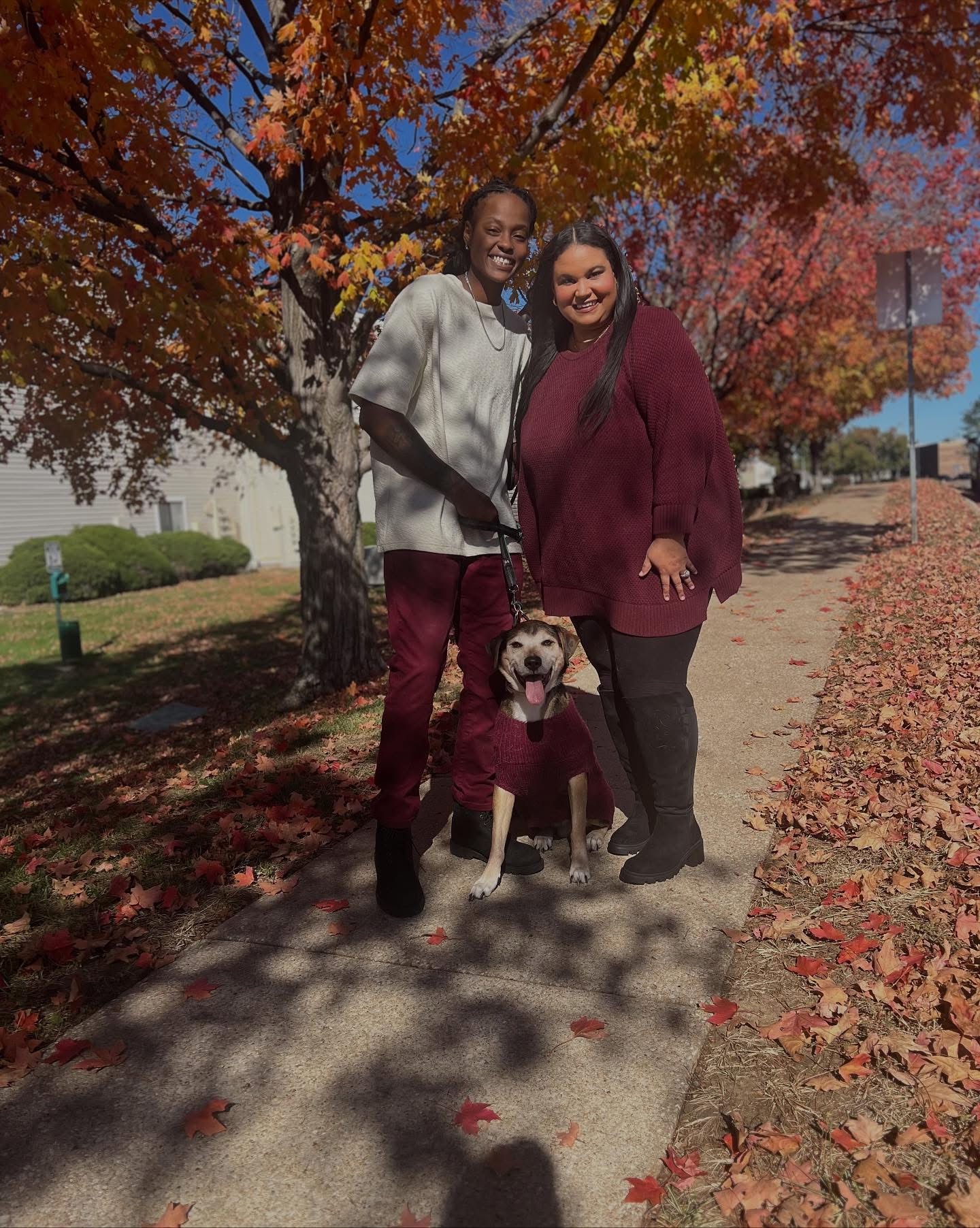 Two women and a dog standing on a sidewalk with autumn trees and fallen leaves, smiling at the camera.