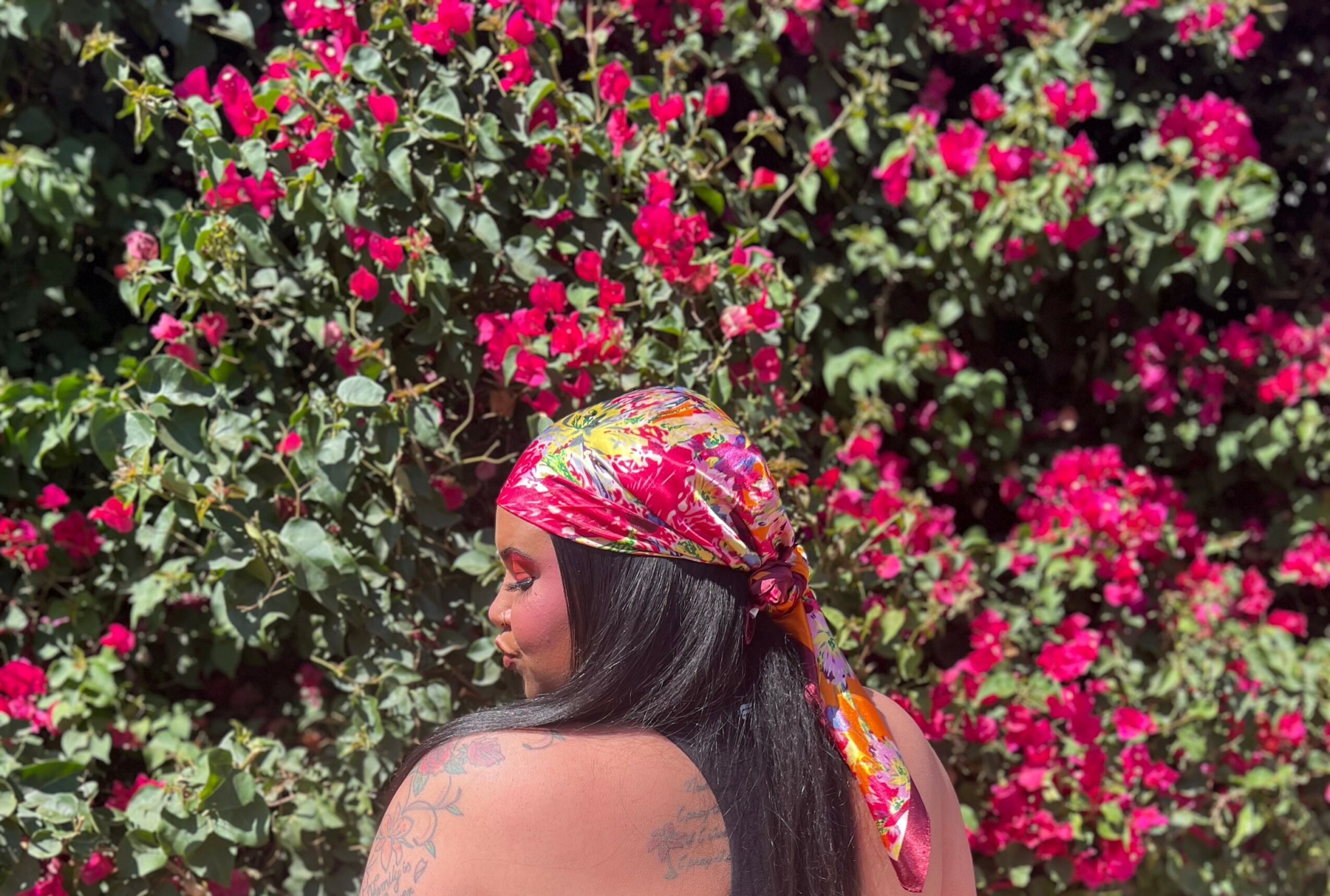 Woman with long dark hair wearing a colorful headscarf, standing in front of pink and red flowers, looking to the side.
