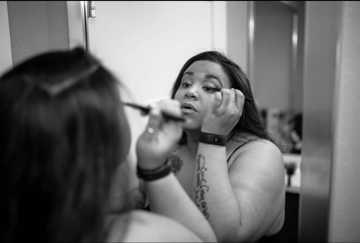 Woman applying makeup while looking in a mirror, with her hand resting on her face, in black and white.