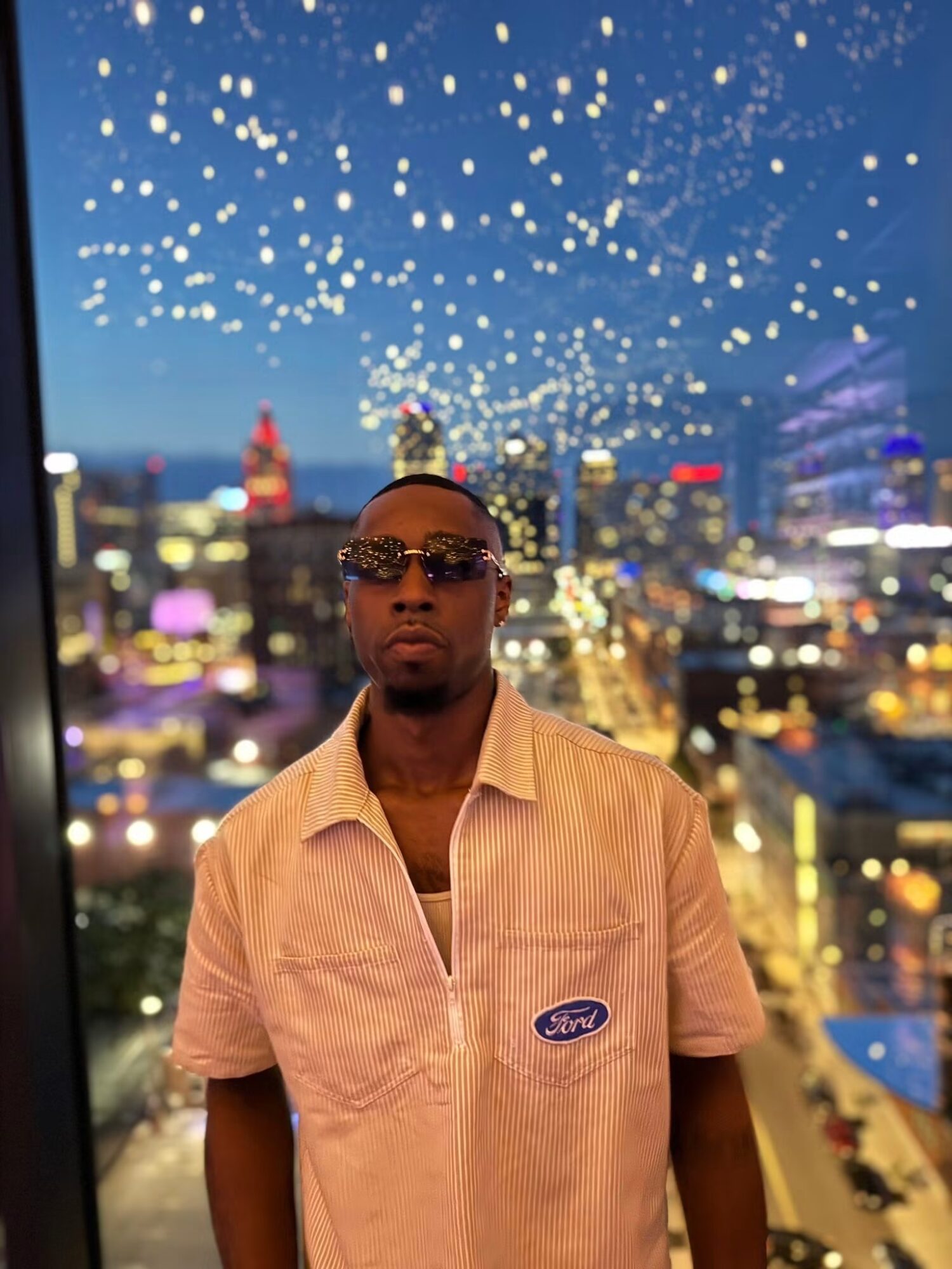 Man wearing sunglasses and a beige shirt with a Ford logo, cityscape with lights and fireworks in background.