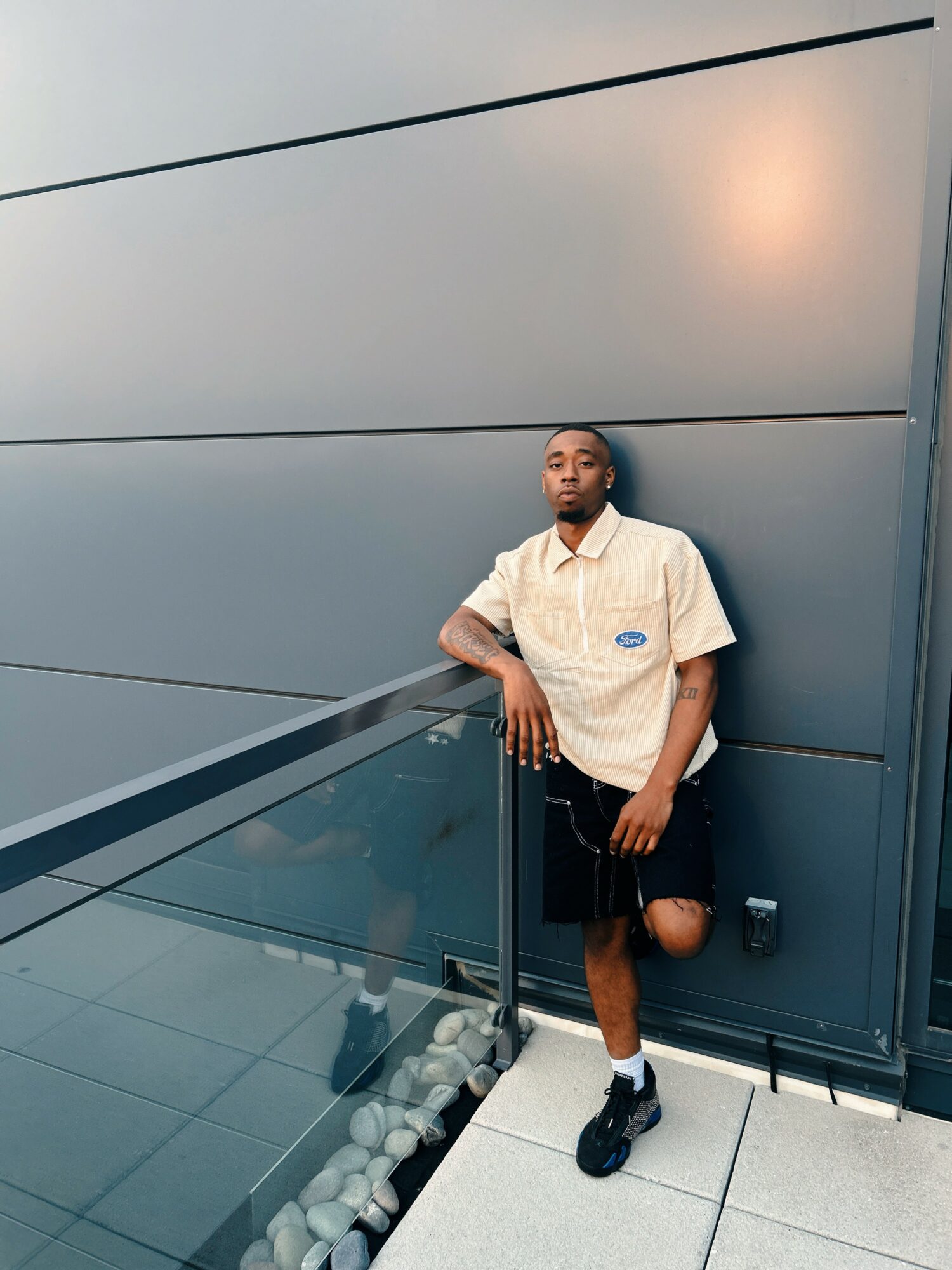 Young man leaning against a wall with a glass railing, wearing a white shirt, shorts, and sneakers.