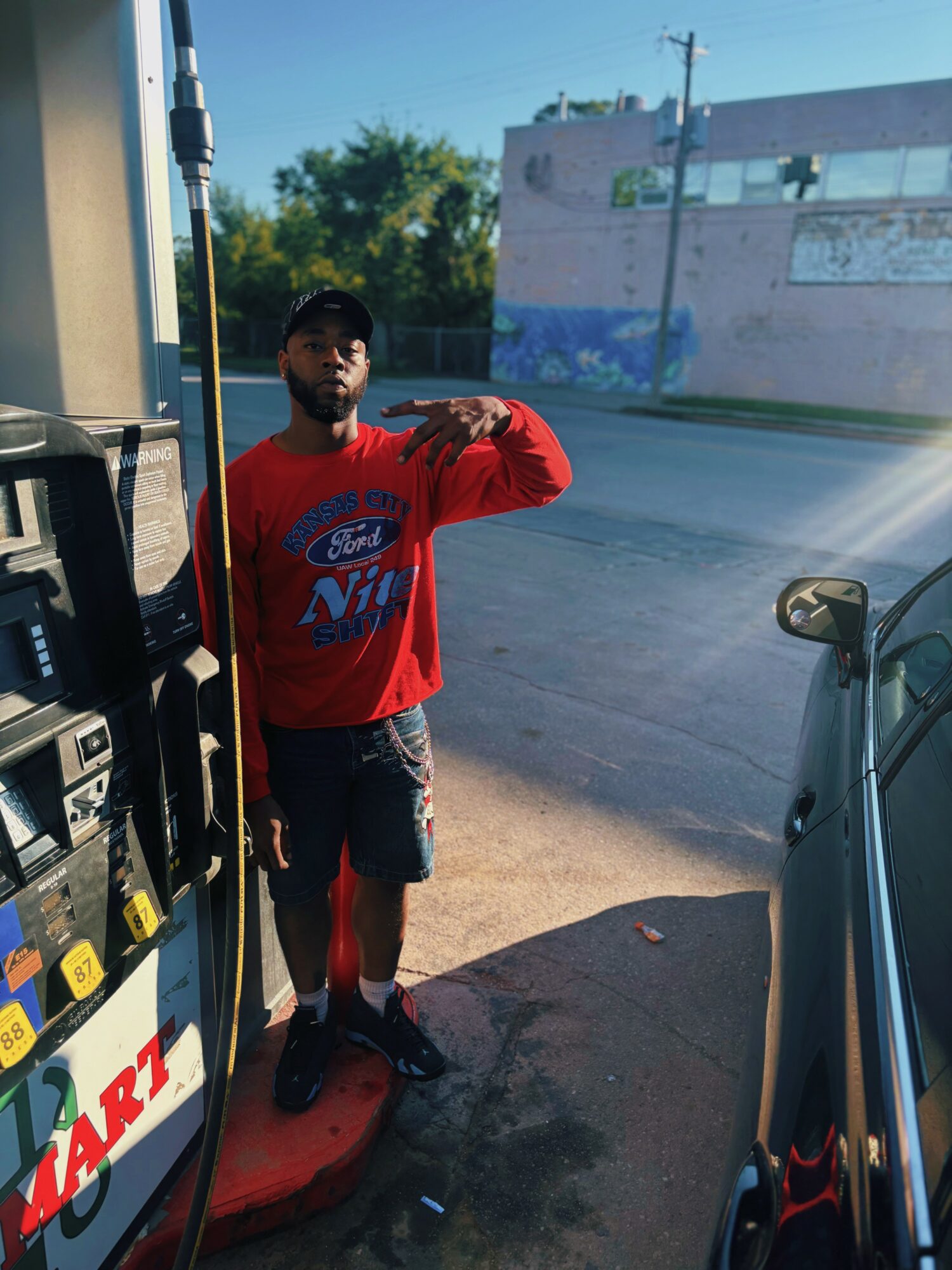 Person taking a mirror selfie outdoors, wearing a red long-sleeve shirt and dark shorts, standing on a sidewalk near a parked car.