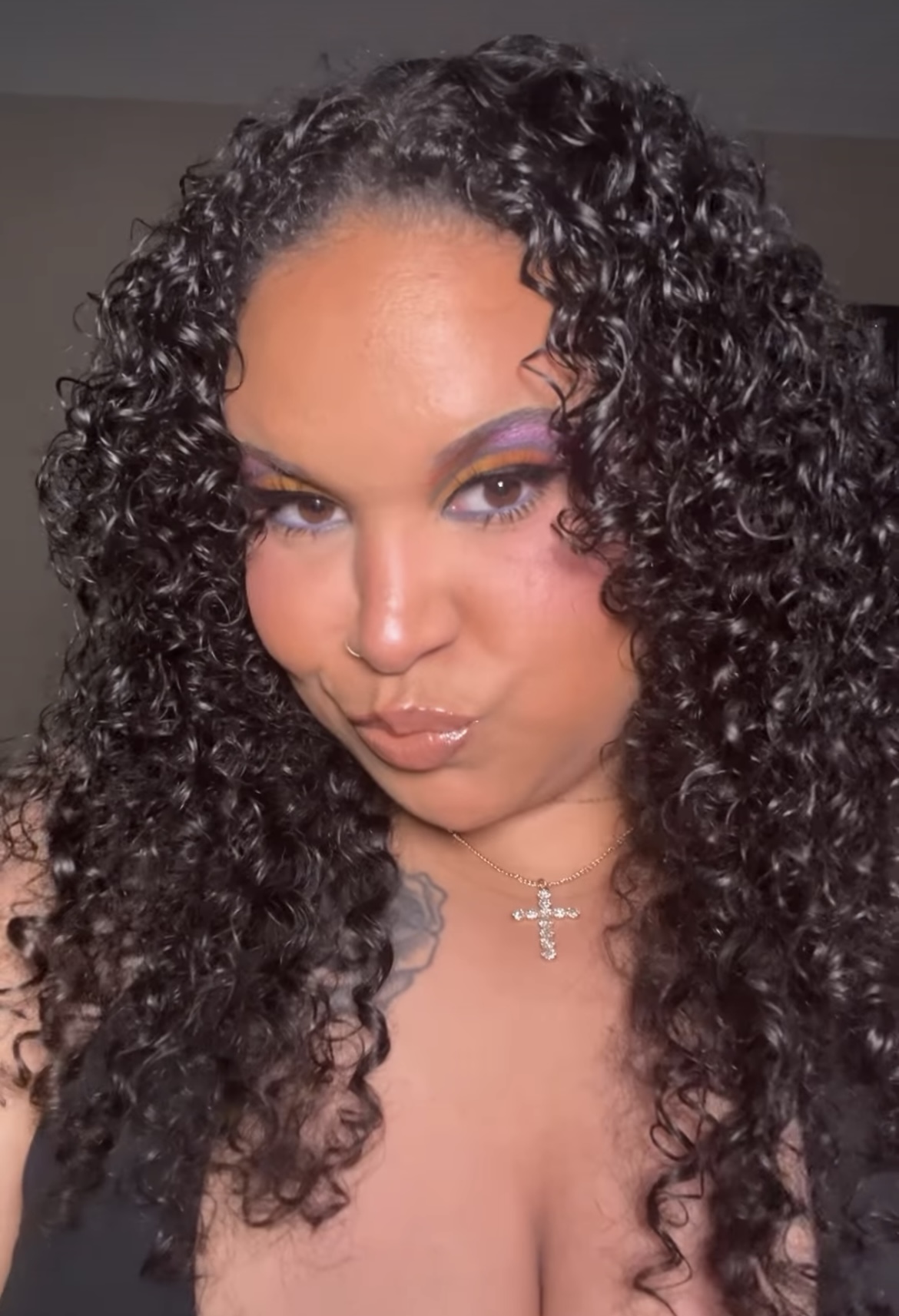 Close-up of a woman with curly hair, makeup, and a necklace, looking at the camera.