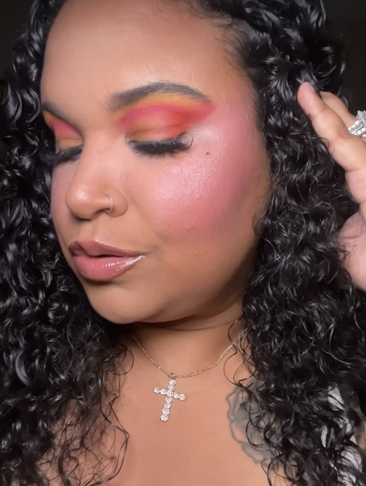 Woman with curly hair, colorful eye makeup, and a cross necklace, looking down with closed eyes.