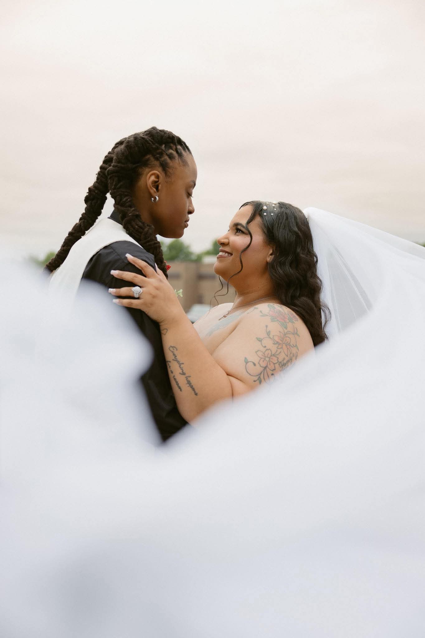 Two women, one with braided hair and the other with dark hair and a veil, look at each other closely outdoors.