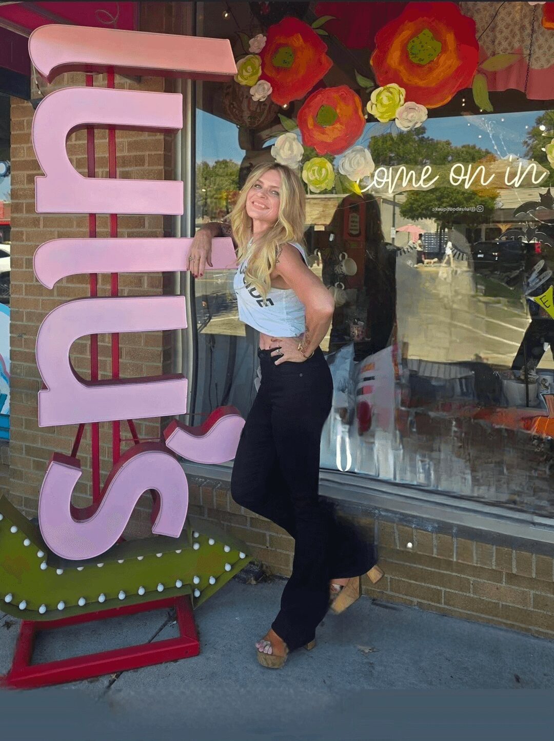Young woman with blonde hair and glasses standing outside a shop, smiling, with large pink letters spelling 'SUE' and colorful flowers in the background.