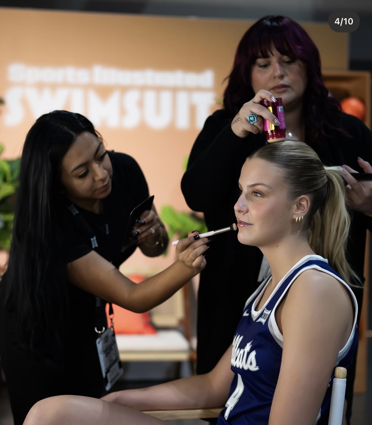 A young woman in a sports jersey gets makeup applied by two women, one holding a brush and the other with a bottle, in a studio setting.