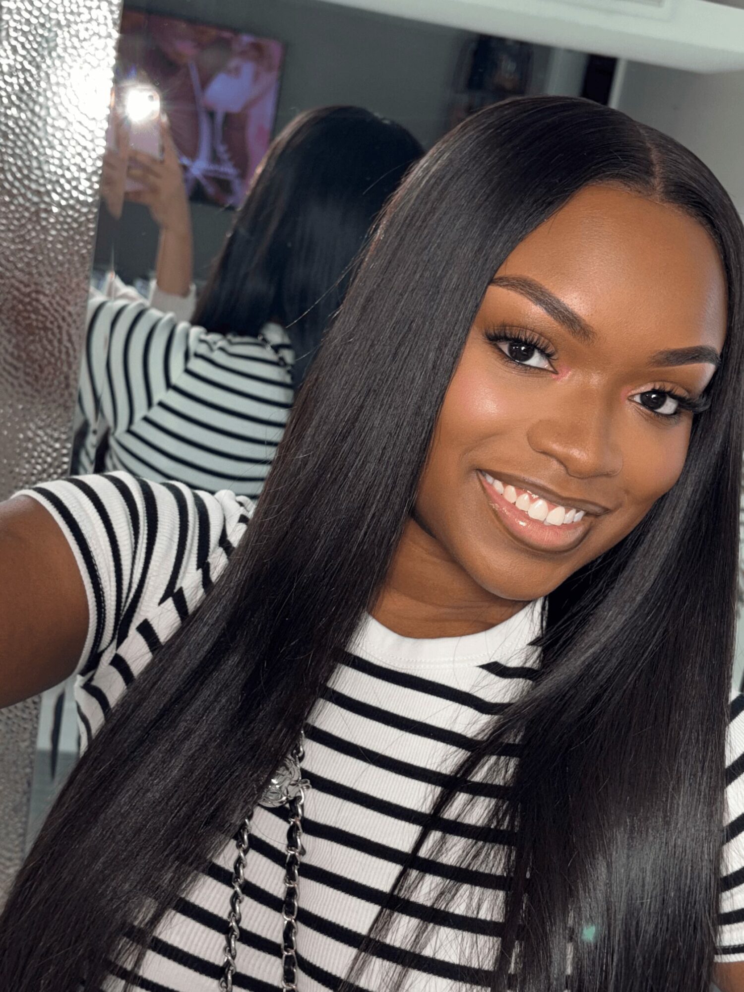 Young woman with long straight hair smiling, wearing a striped shirt, mirror reflection behind her.