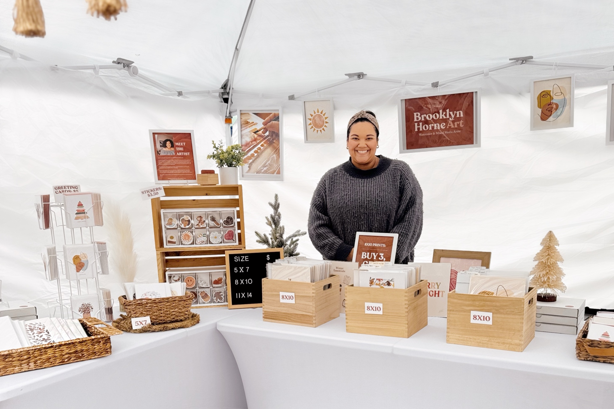 Smiling person standing behind a table with boxes and holiday decorations inside a white tent.