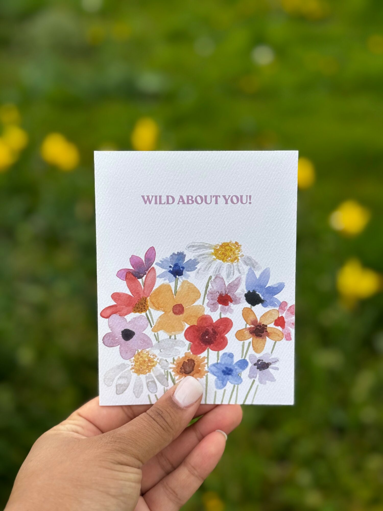 Hand holding a card with colorful flowers and the text 'WILD ABOUT YOU!' against a blurred green and yellow background.