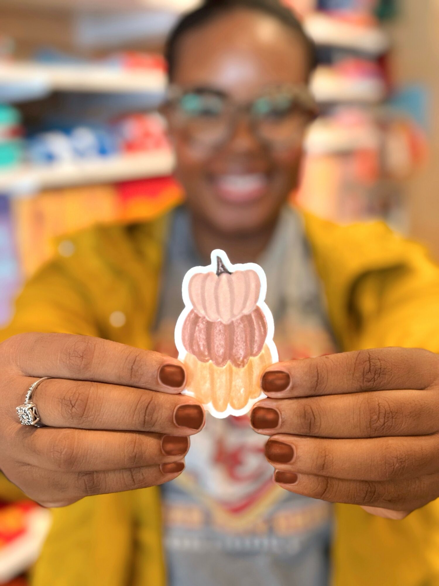 Person holding a sticker of a pumpkin with pink and orange stripes, smiling in a store, wearing glasses and a yellow jacket.