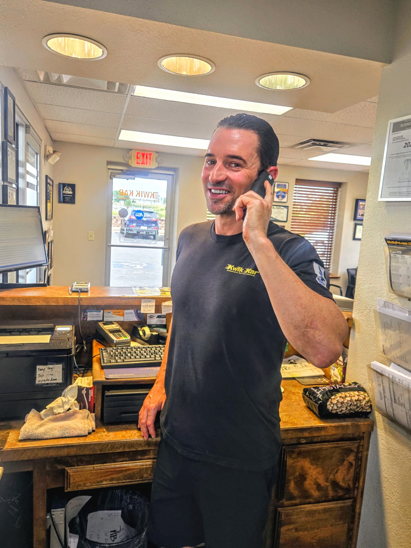 Man smiling and talking on a phone inside a store, standing near a wooden counter with various items.