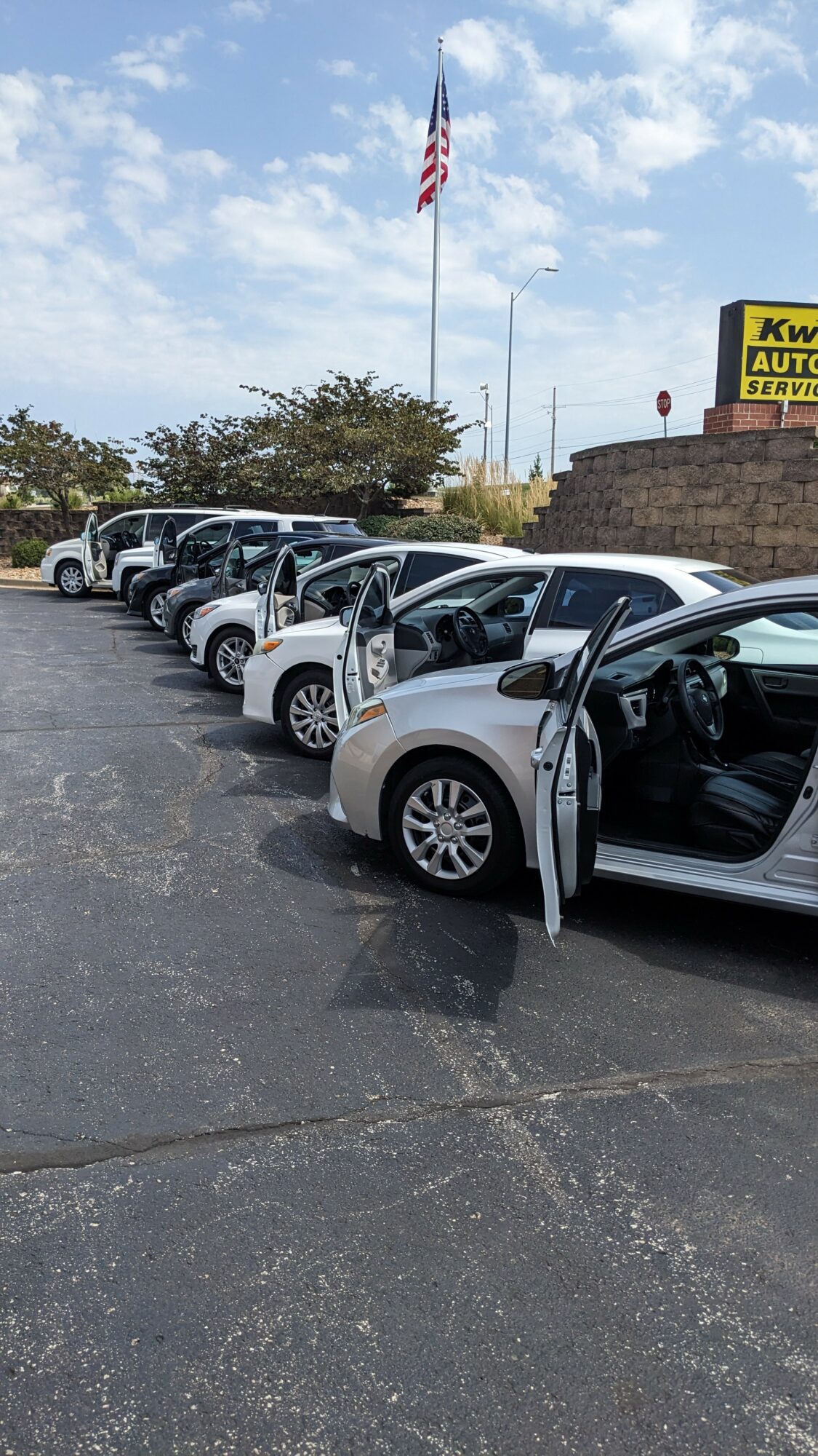 Line of white cars parked with open doors, American flag flying in the background, cloudy sky above.