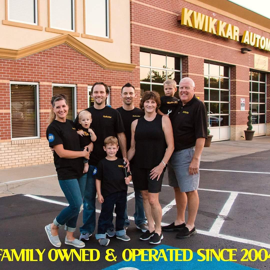 Group of seven people, including children, standing outside a building with a sign reading 'KWIK KAR AUTO.' They are smiling and posing for the photo.