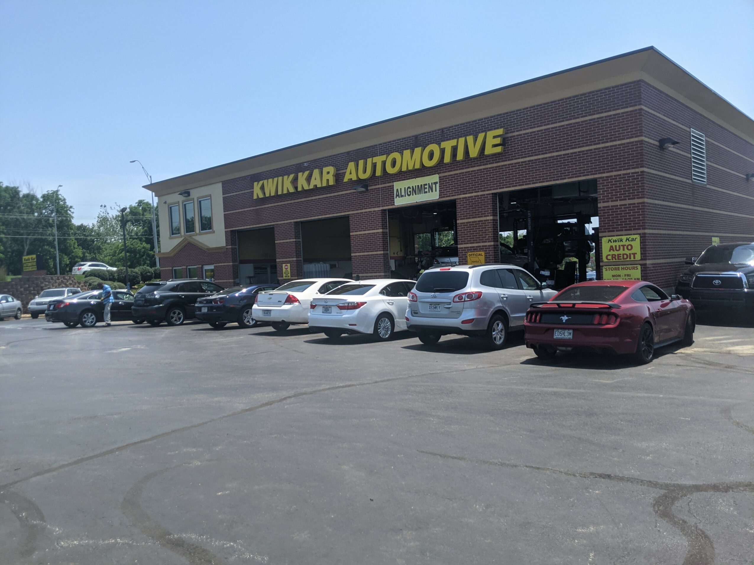 Car dealership building with cars parked in front, sign reads 'KWIK KAR AUTOMOTIVE', clear blue sky, parking lot.