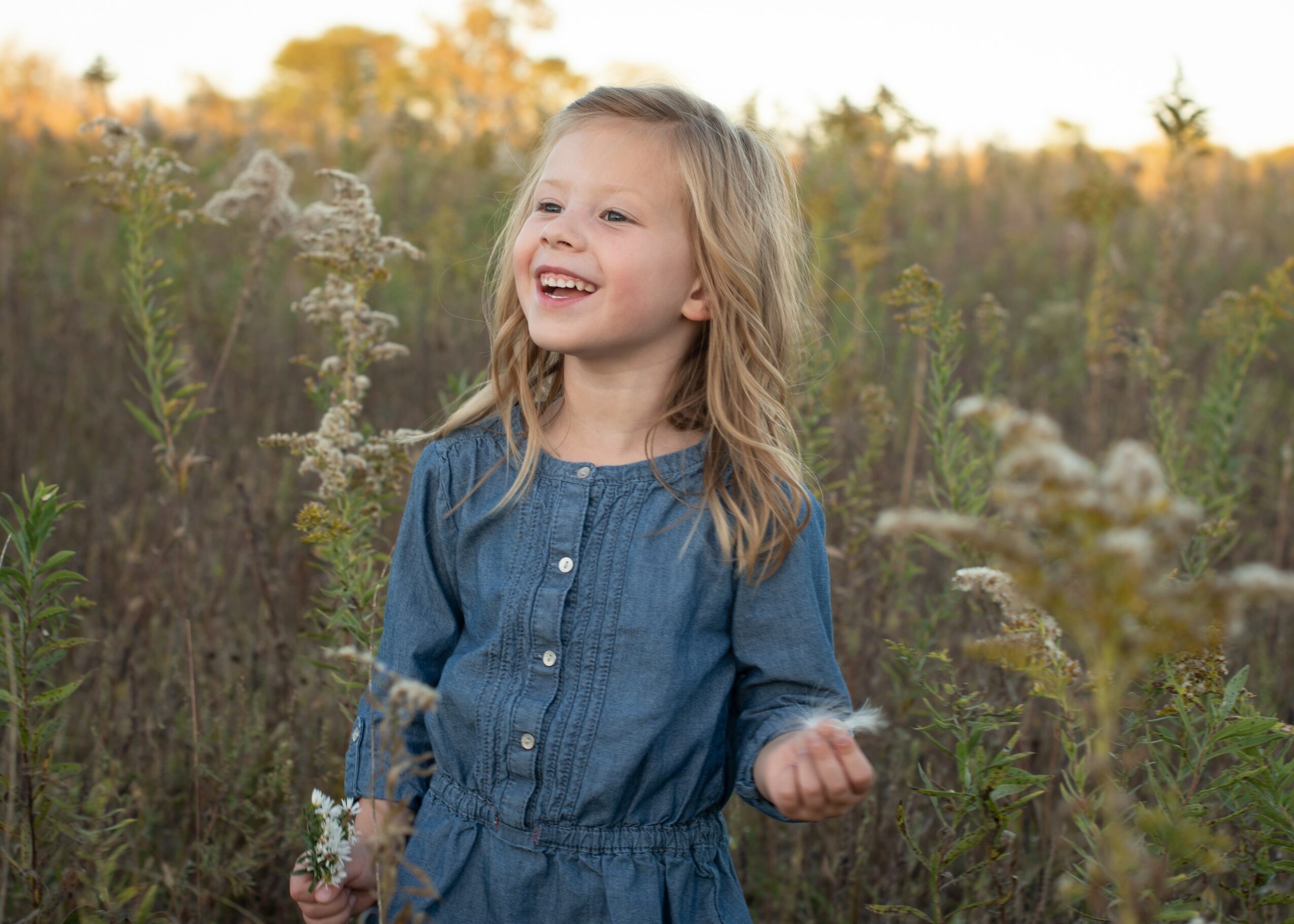 Young girl with blonde hair smiling in a field of tall plants and wildflowers during sunset.