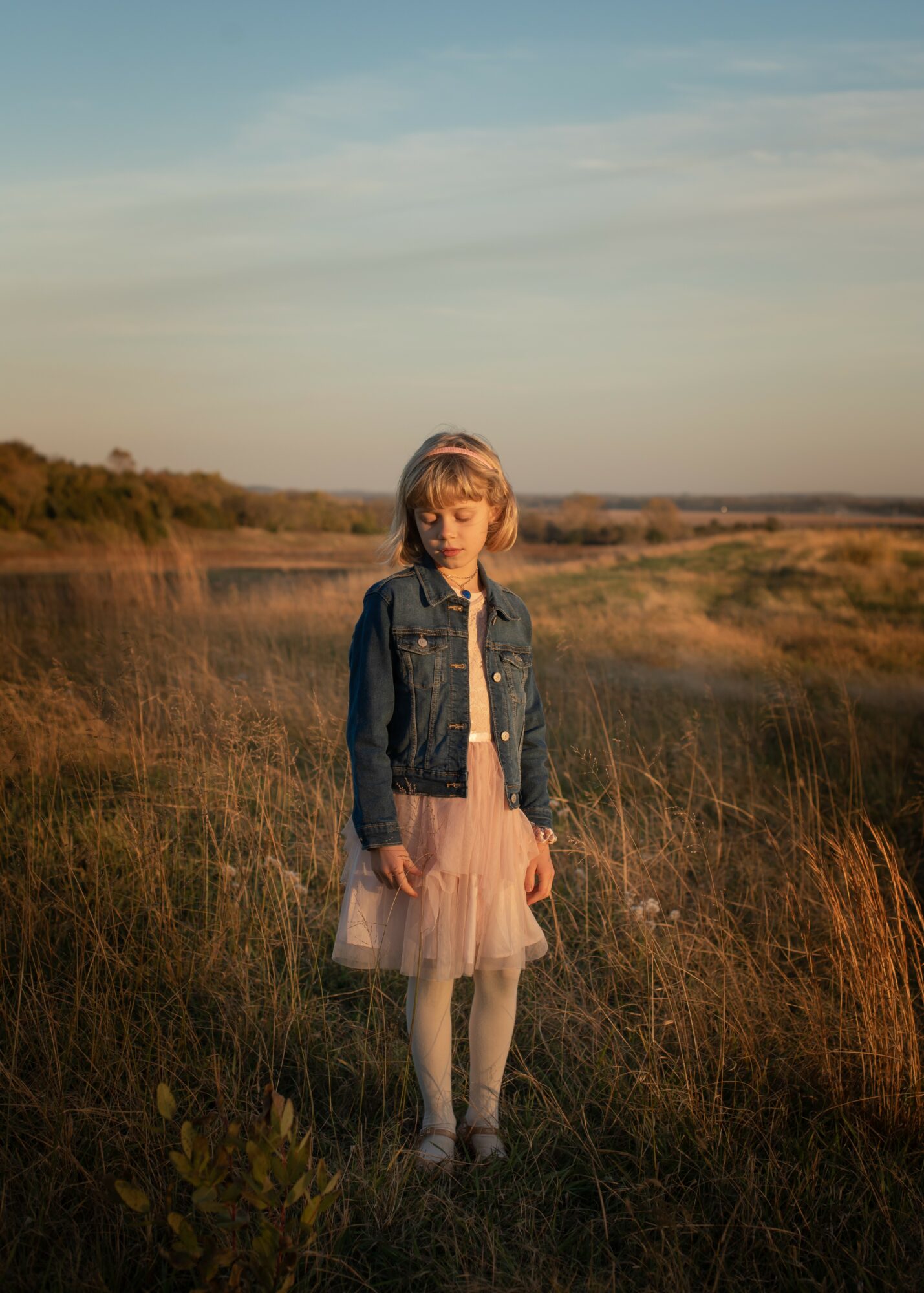 Girl standing in a field with tall grass, wearing a denim jacket and pink dress, under a partly cloudy sky.