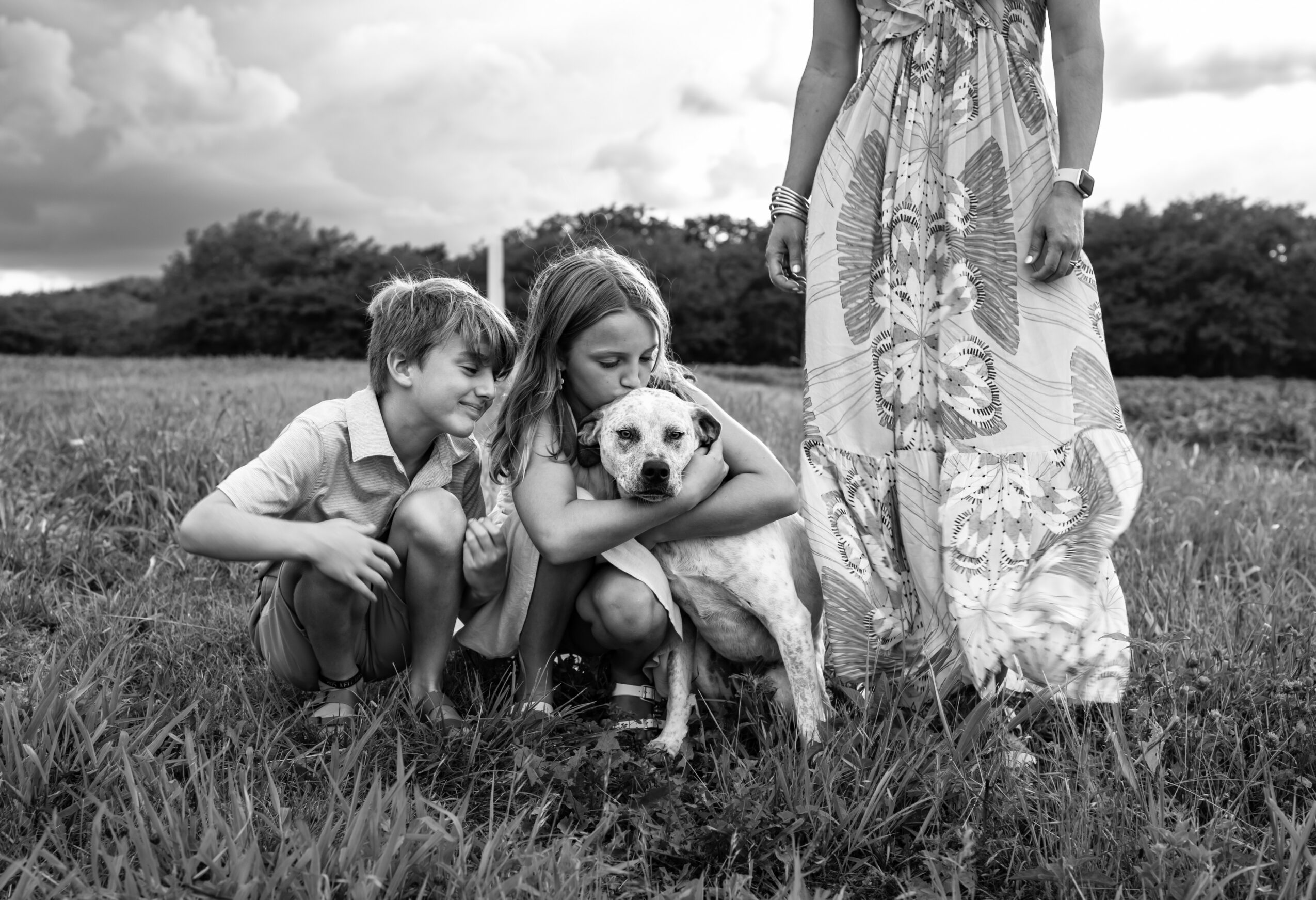 Two children and a dog in a grassy field with a person standing nearby, cloudy sky in background.