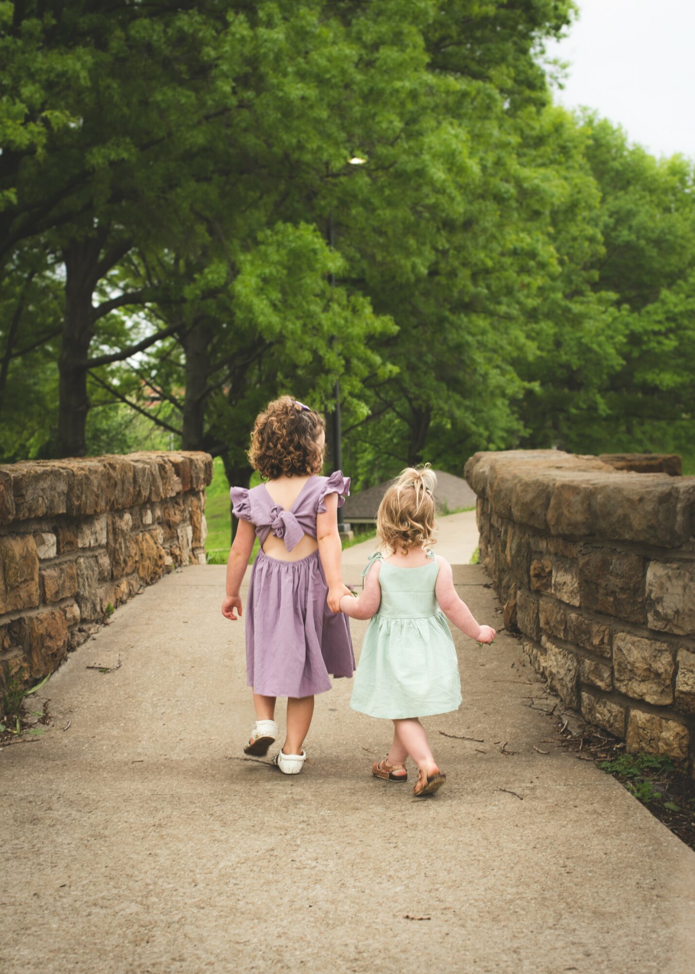 Two young girls walk hand in hand on a stone bridge with green trees in the background.