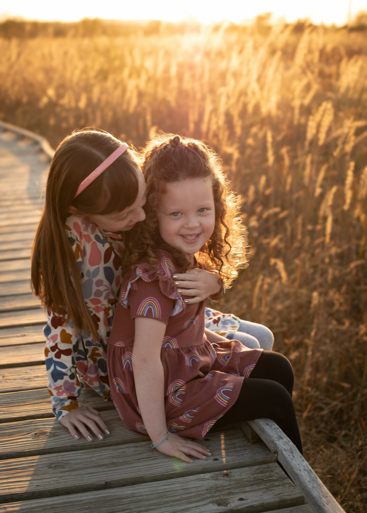 Two young girls sitting on a wooden boardwalk in a field during sunset, smiling and hugging each other.
