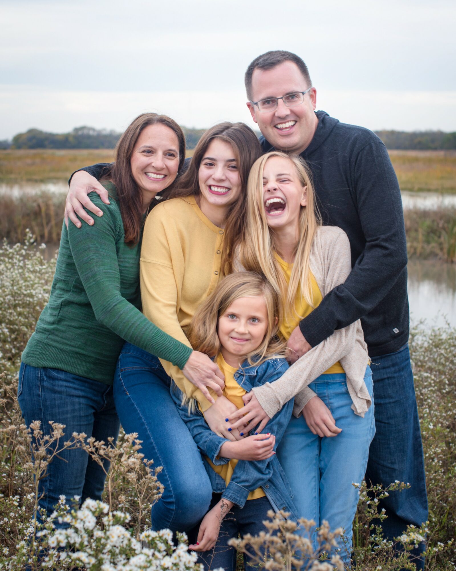 Group of five people smiling outdoors, standing close together with arms around each other, in a natural setting with water and grass.