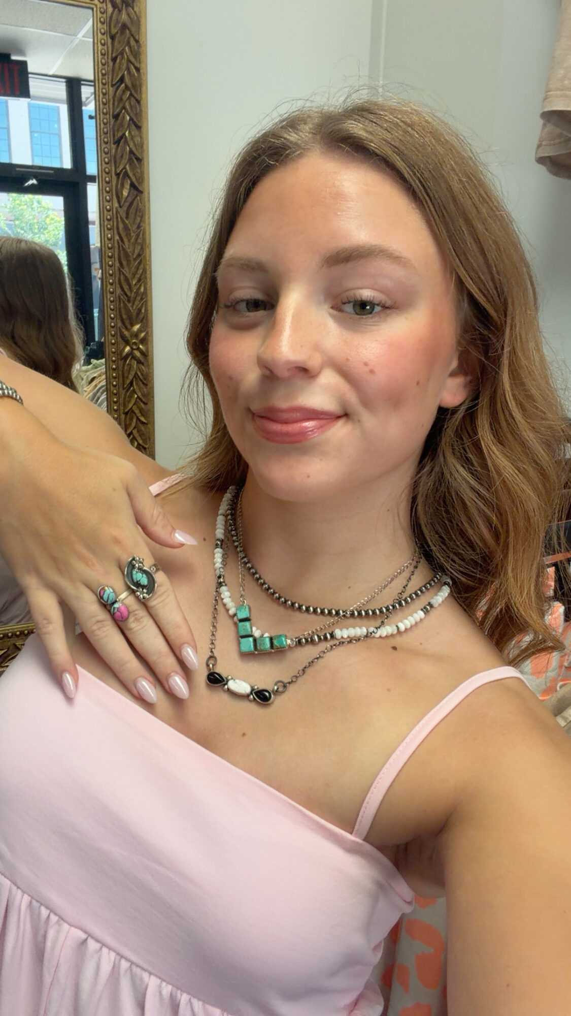 Young woman with light brown hair wearing layered necklaces and a pink top, smiling softly, in front of a mirror.