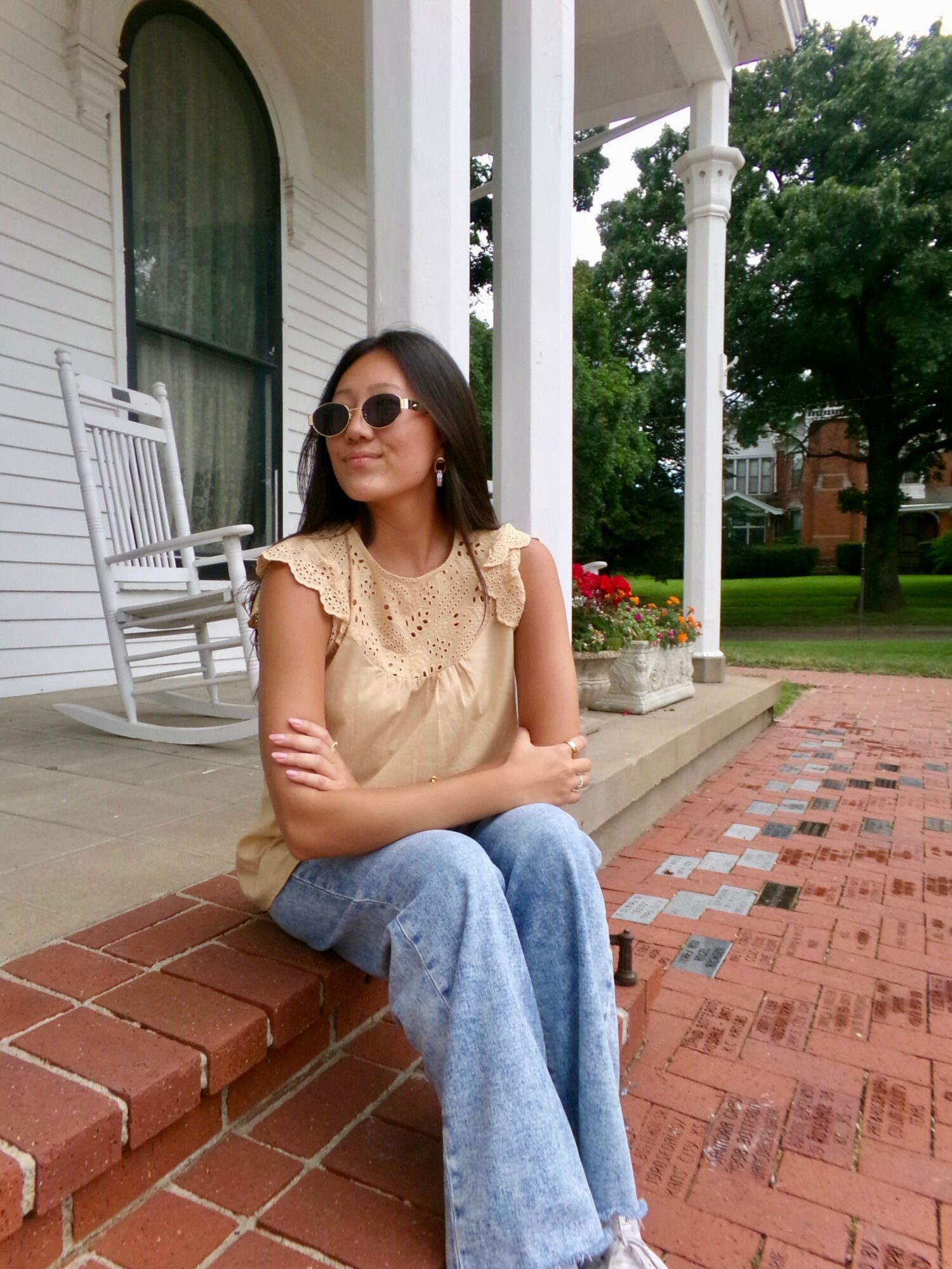 Woman sitting on brick steps outside a house, wearing sunglasses, a beige top, and jeans, with greenery and a porch in background.