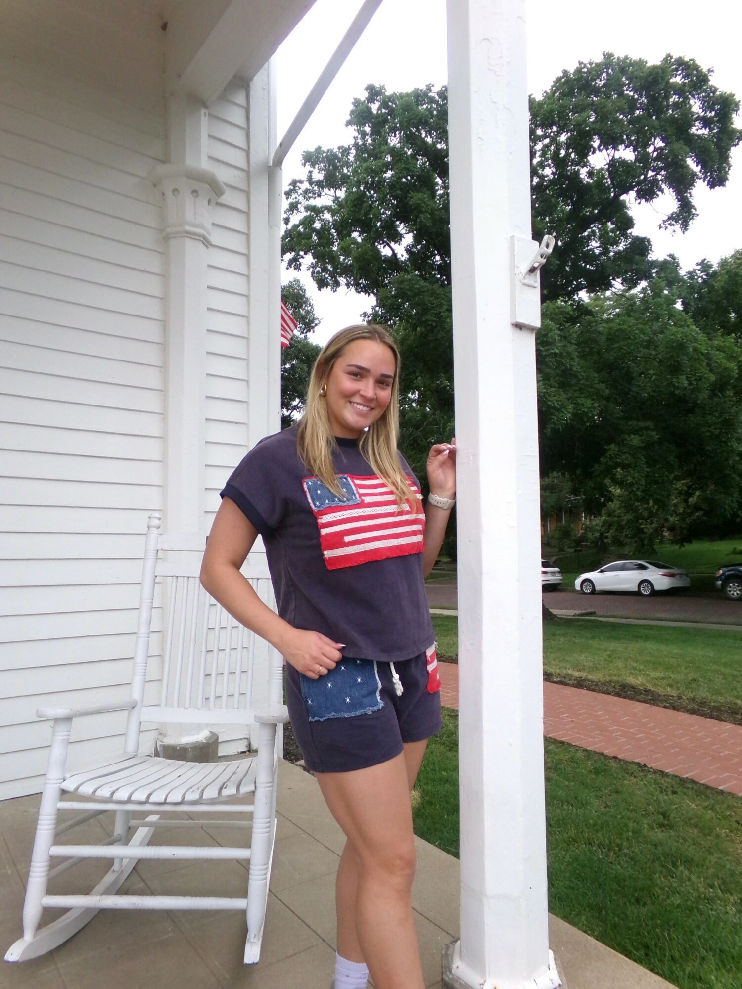 Young woman with long hair smiling outdoors, standing next to a white porch column, wearing a navy T-shirt and shorts.
