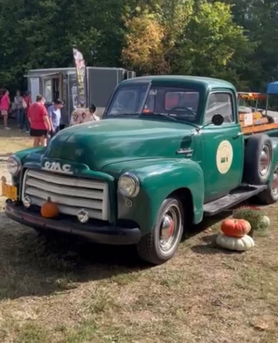 Green vintage truck with pumpkins around it, people and trees in the background, outdoor setting, sunny day.