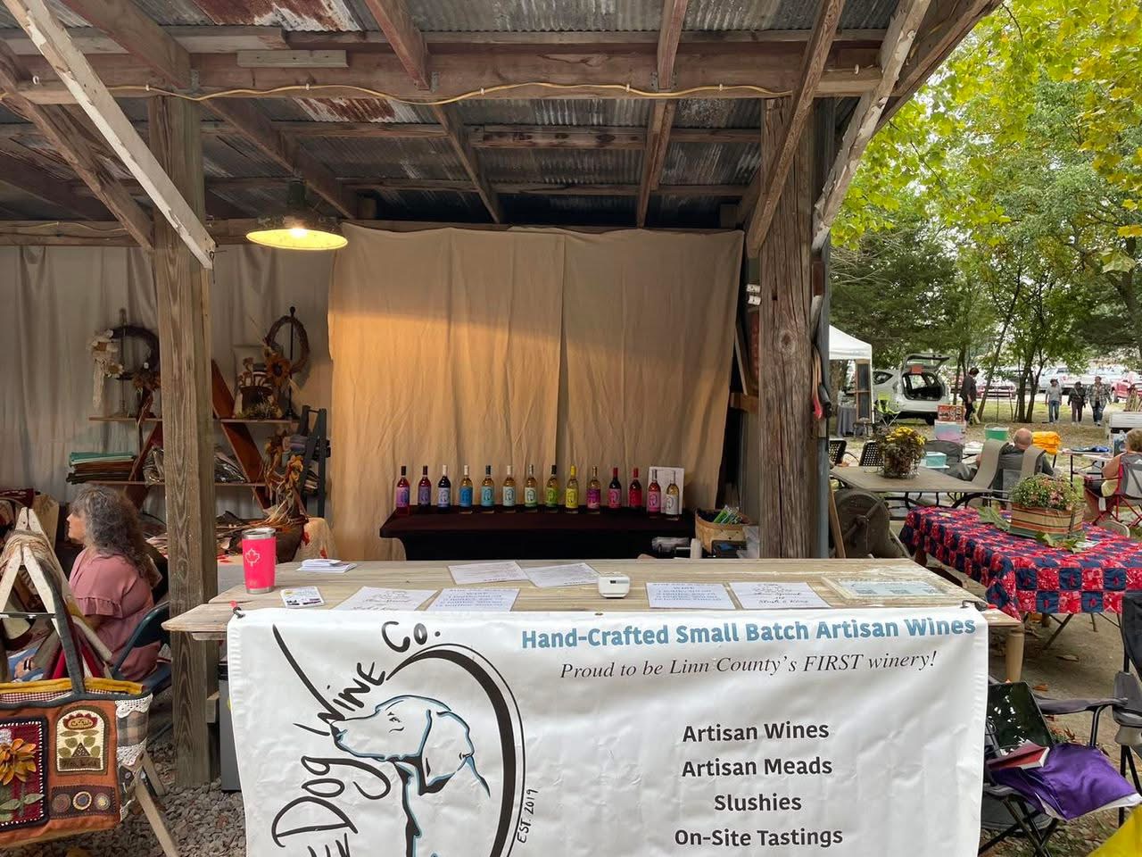 Indoor market stall with bottles of wine, shelves, and a curtain backdrop, surrounded by tables and outdoor trees.