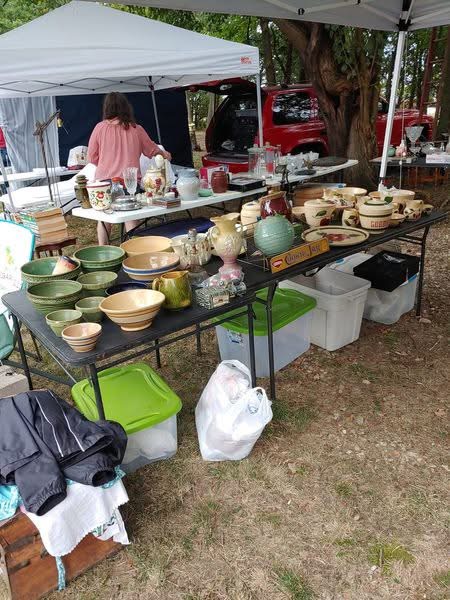 Outdoor market stall with various ceramic bowls, vases, and kitchenware on a black table, with tents and trees in background.