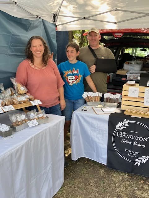 Three people standing behind bakery tables at an outdoor market, with baked goods and a sign for Hamilton Christian Bakery.
