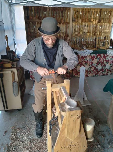 Man wearing a hat and glasses carving wood at a workshop with tools and wooden utensils displayed behind him.