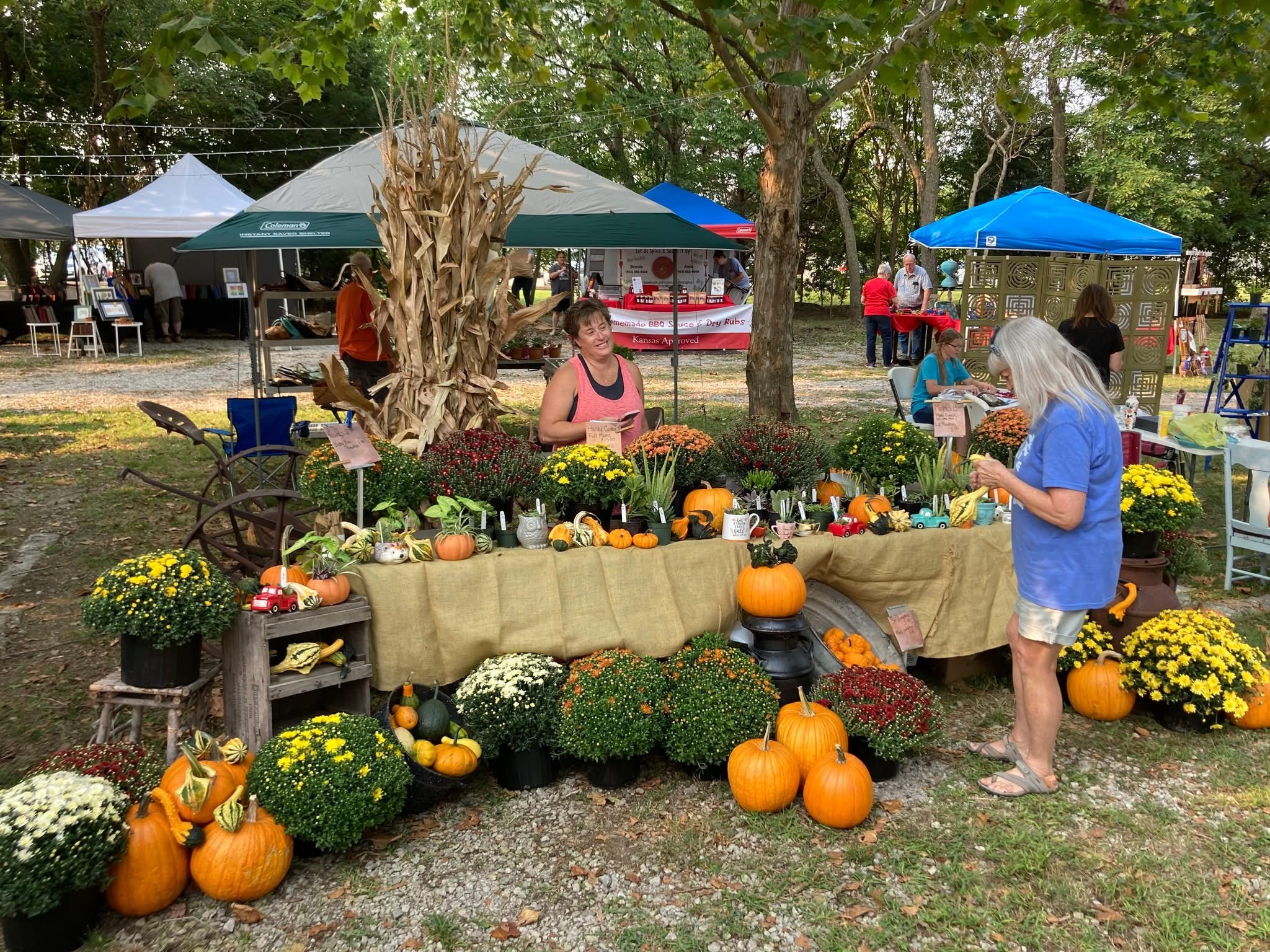 People shopping at a pumpkin and flower stand outdoors with tents and trees in the background.