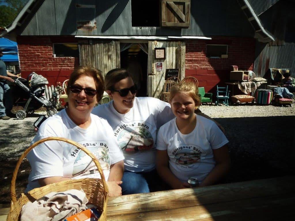 Three people smiling outdoors in front of a rustic building, wearing white shirts, with bags and chairs nearby.