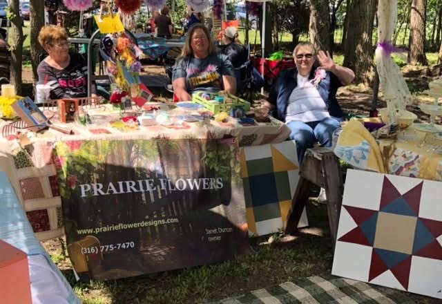People sitting at a table with colorful crafts and flowers outdoors, with a sign for Prairie Flowers in front.