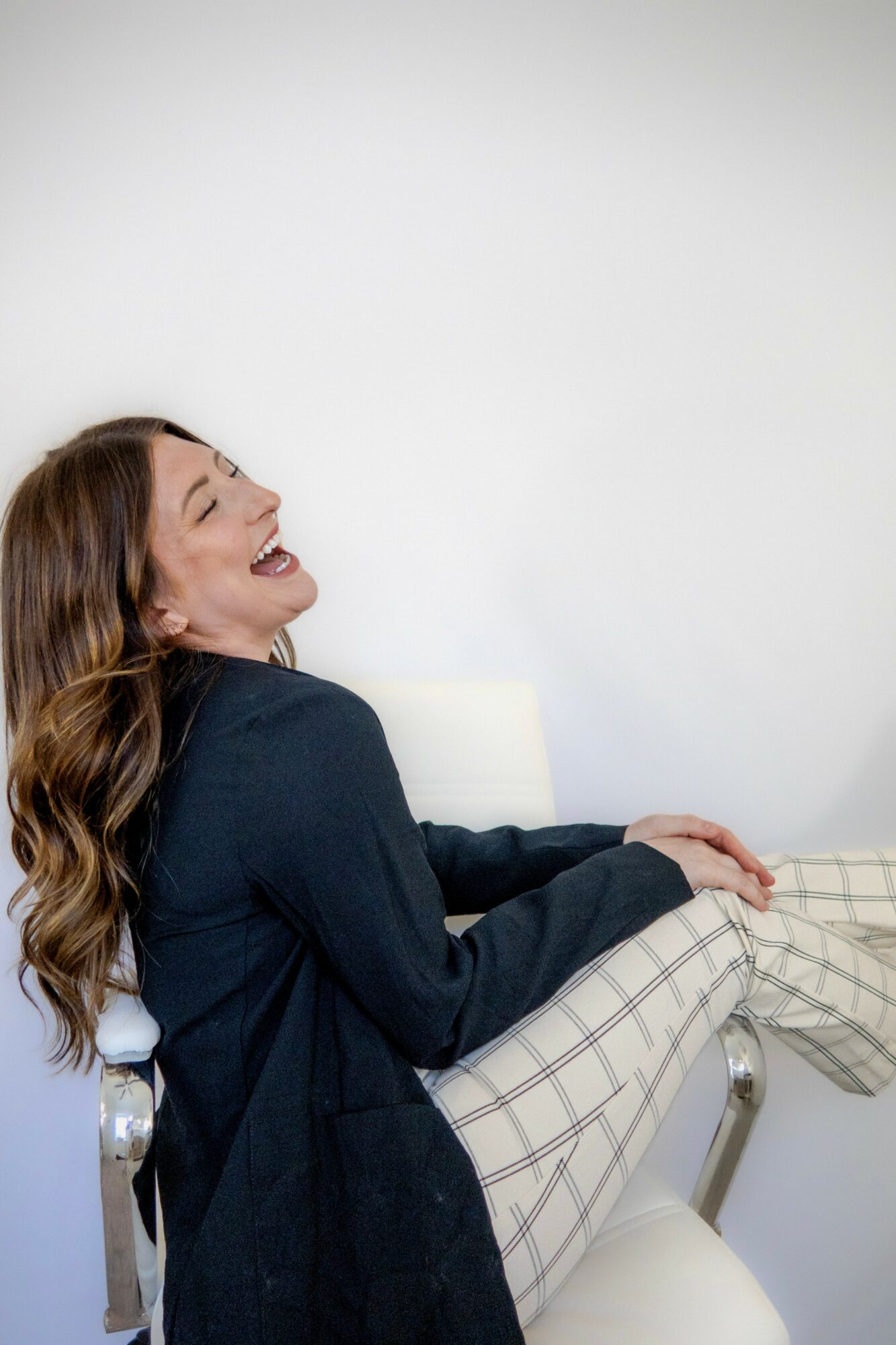 Woman with long hair laughing, sitting on a chair with a plain white wall background.