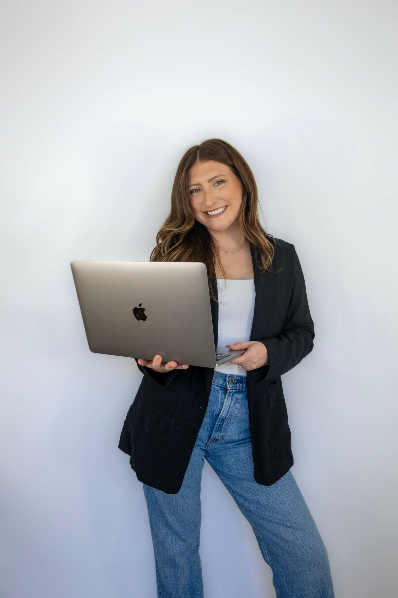 Woman with long hair smiling, holding a laptop, wearing a black blazer and jeans, standing against a white background.