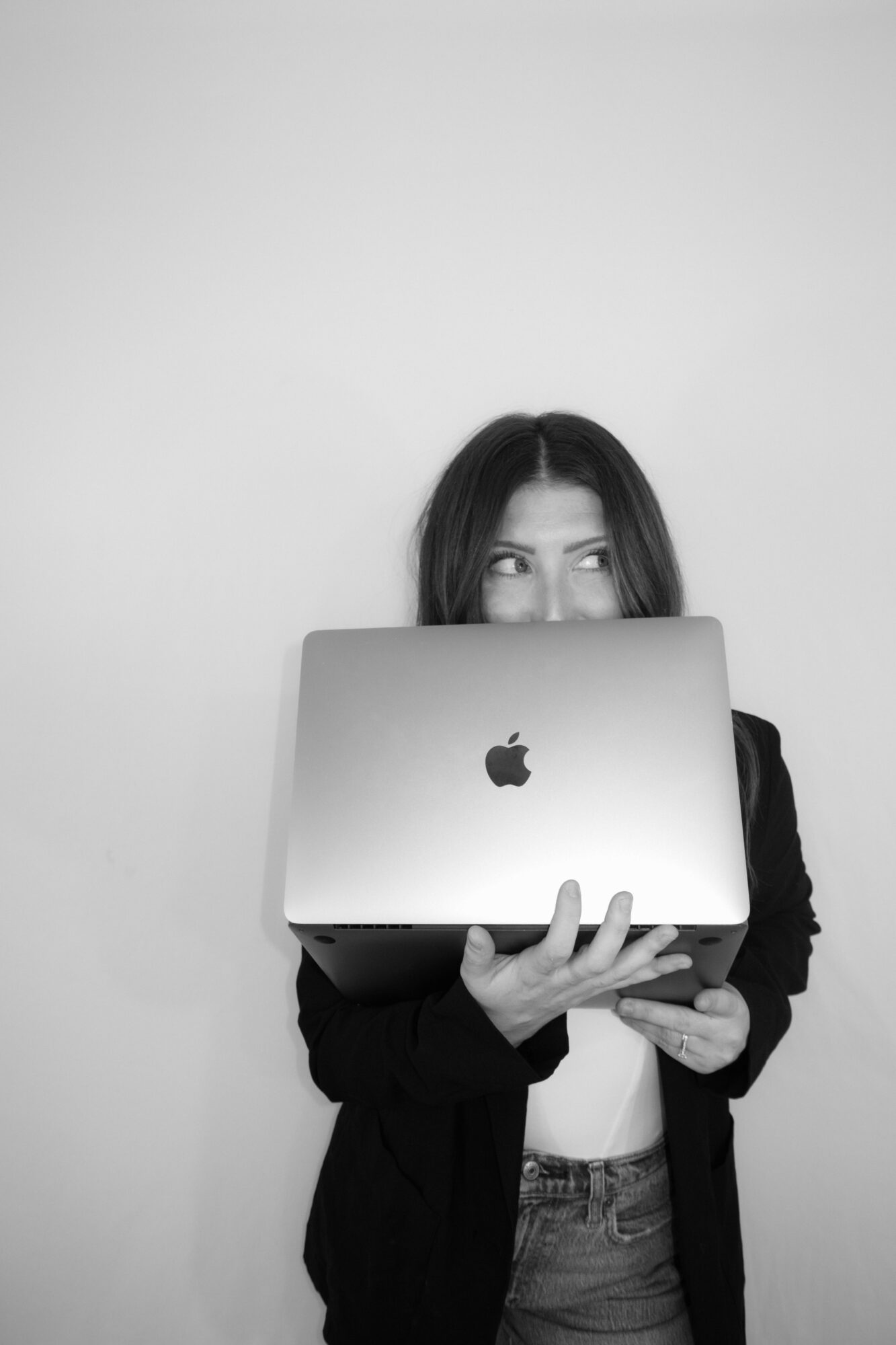 Person holding a laptop in front of face, eyes visible above, against plain background.