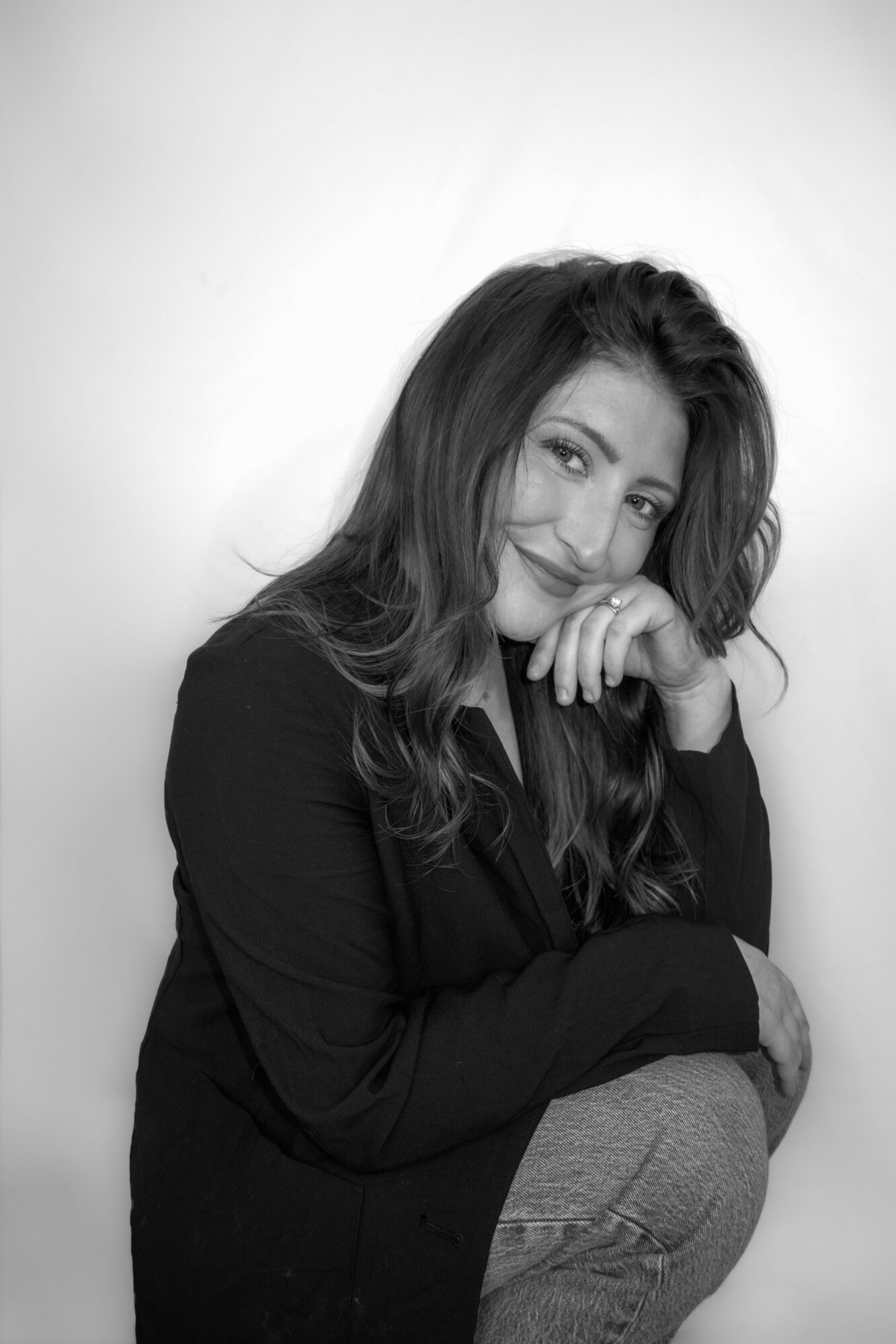 Woman with long hair smiling, resting her chin on her hand, wearing a dark top, against a plain background.