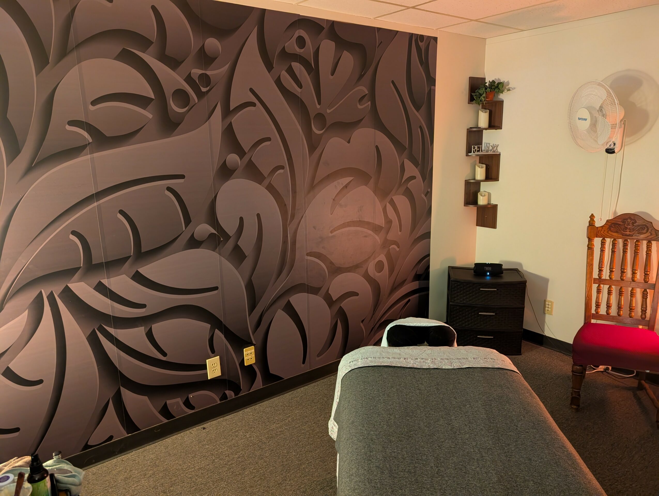 Room with decorative wall, massage table, chair, and small shelf with plants, clock, and furniture.