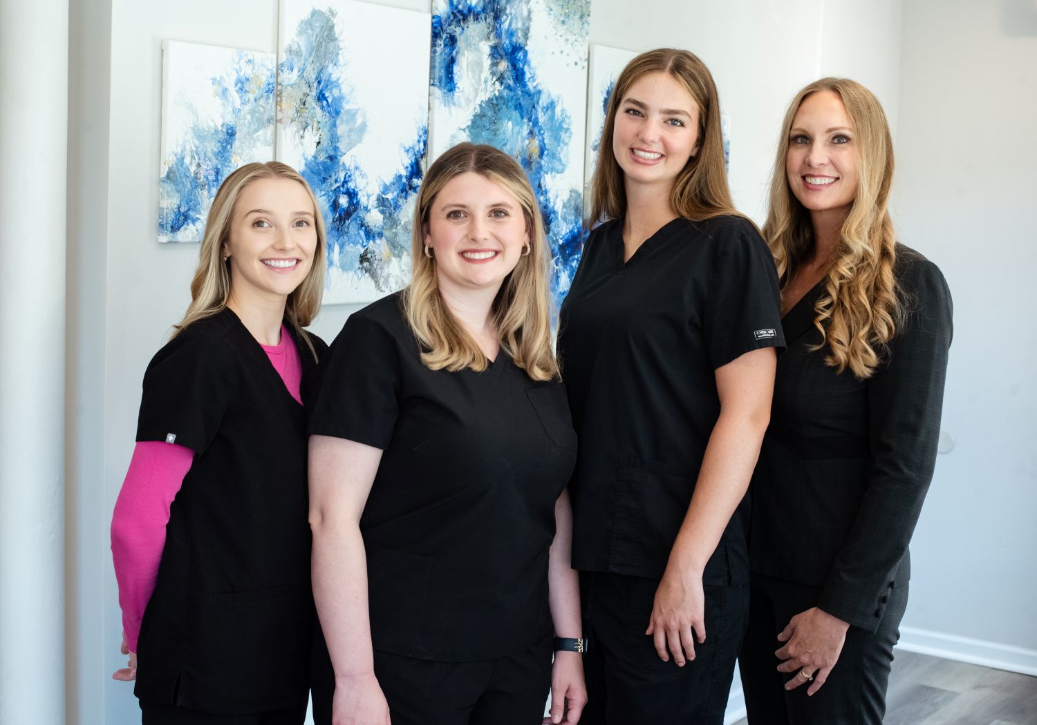 Four women stand together indoors, smiling, with abstract blue and white artwork behind them.